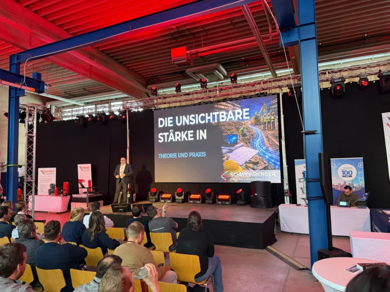 Ein Referent hält vor interessiertem Publikum einen Fachvortrag zum Thema „Die unsichtbare Stärke in Theorie und Praxis“ auf einer großen Event-Bühne mit Firmenlogo. Diese Initiative von Schweinberger Glasfaser Experte fördert das Know-how rund um den Glasfaseranschluss und die Qualitätssicherung.