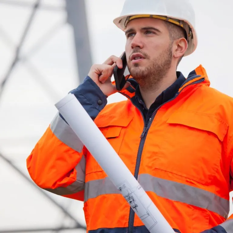 Dieses Bild zeigt einen Arbeiter in orangener Arbeitsjacke und einem weißen Schutzhelm. In der einen Hand hält er einen Plan und mit der anderen telefoniert er mit seinem Smartphone