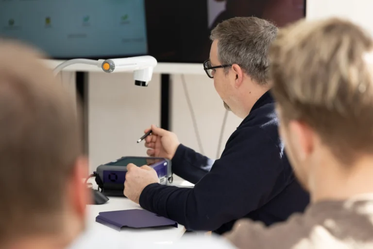 Ein Experte bedient konzentriert ein OTDR-Messgerät während einer Schulung oder Analyse, um die Signalqualität einer Glasfaserstrecke zu prüfen. Im Vordergrund sind die Hinterköpfe von Teilnehmern zu sehen, die den Prozess an einem Schreibtisch mit technischem Equipment verfolgen.